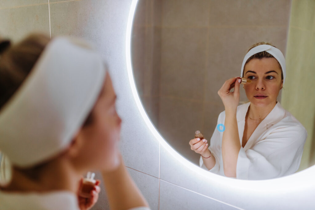 Nahaufnahme einer jungen Frau, die sich im Badezimmer Kosmetiköl aus einer Flasche ins Gesicht aufträgt. Spiegelbildaufnahme.