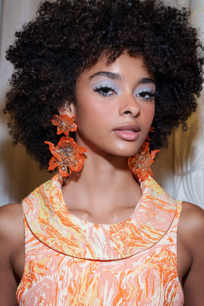 Model mit pastellfarben geschminkten Augen, Korallenohrringen und organgenem Oberteil, Style Costelloe