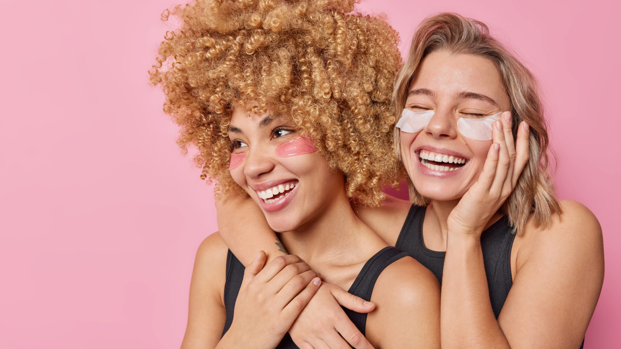 Zwei lebensfrohe Frauen pflegen ein freundschaftliches Verhältnis, umarmen sich, lächeln breit, kleben sich Augenpads auf.