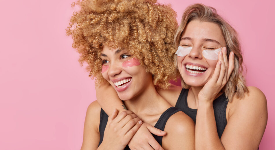 Zwei lebensfrohe Frauen pflegen ein freundschaftliches Verhältnis, umarmen sich, lächeln breit, kleben sich Augenpads auf.