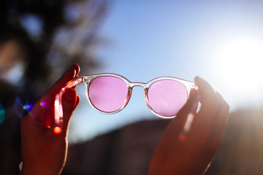 Sommerliches Straßenbild einer Frau mit pinkfarbener Sonnenbrille vor blauem Himmel und Sonne.
