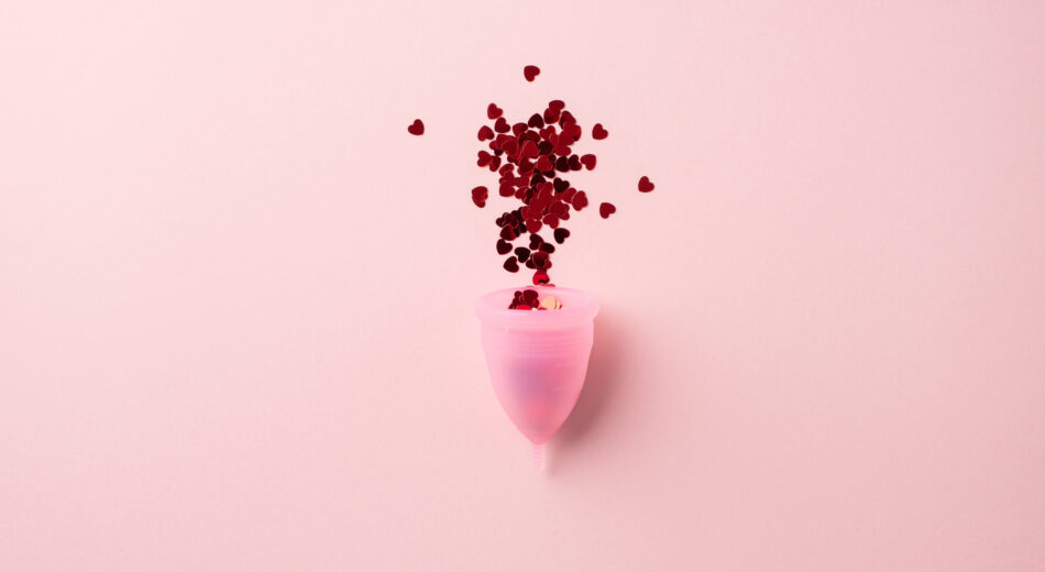 Draufsicht auf eine Explosion roter, herzförmiger Konfetti aus einer rosa Menstruationstasse vor isoliertem pastellrosa Hintergrund