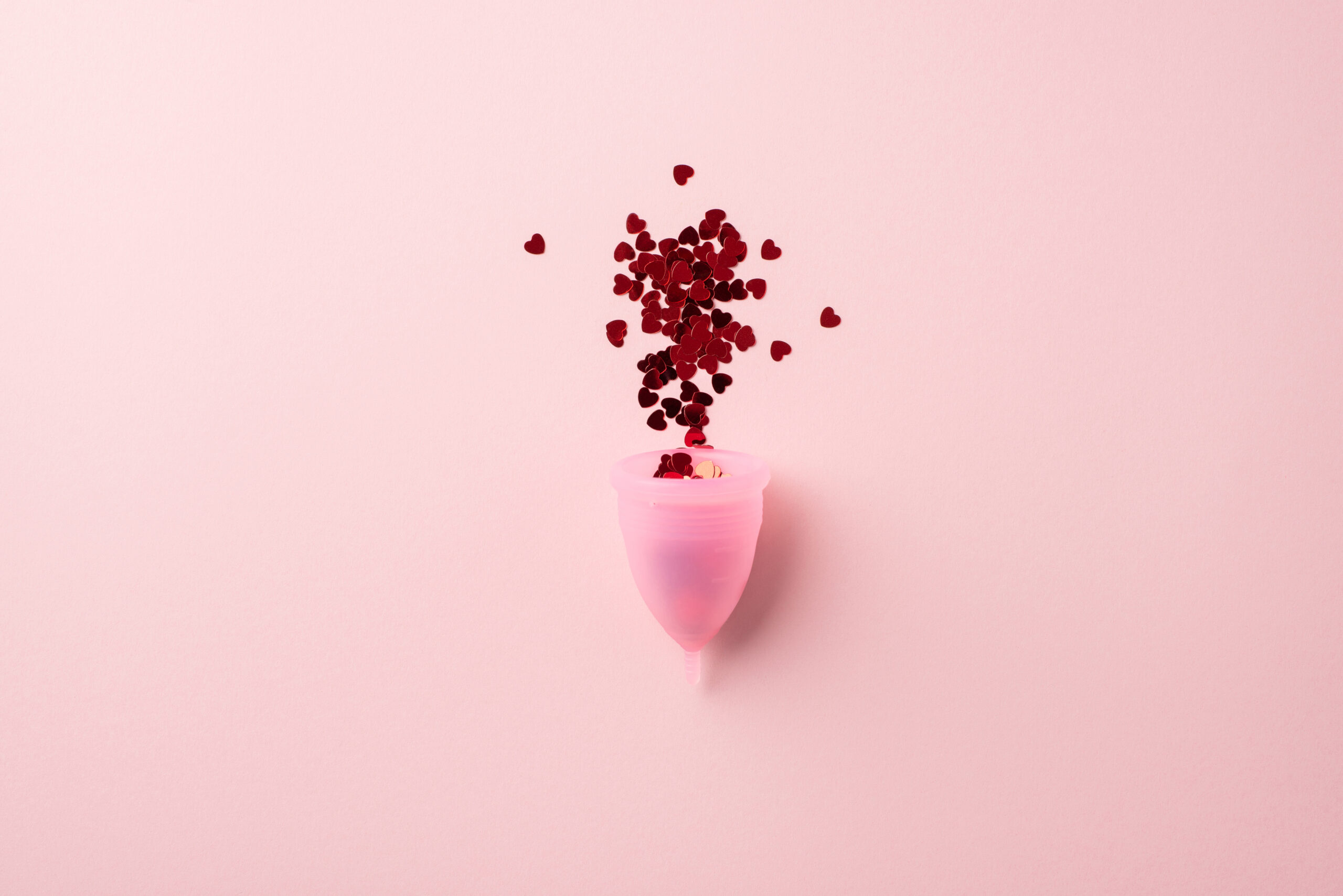 Draufsicht auf eine Explosion roter, herzförmiger Konfetti aus einer rosa Menstruationstasse vor isoliertem pastellrosa Hintergrund