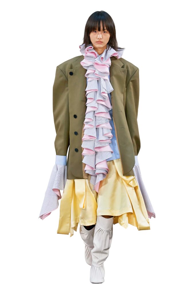 Model am Laufsteg, oversized Blazer in grün, dazu Bluse mit XL Rüschen und gelbem Rock, Kollektion Abra